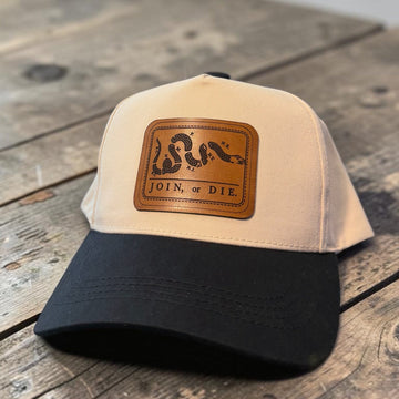 A two-tone trucker cap with a beige front panel and black brim, featuring a brown leather patch on the front with a coiled snake and the words 'JOIN OR DIE'. The hat is photographed on a rustic wooden surface with a blurred gray background.